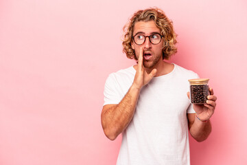 Young caucasian man holding coffee jar isolated on pink background is saying a secret hot braking...