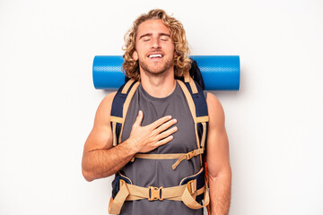 Young mountaineer caucasian man with a big backpack isolated on white background laughs out loudly keeping hand on chest.