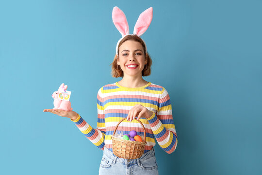 Beautiful young woman with bunny ears, toy rabbit and Easter basket on blue background
