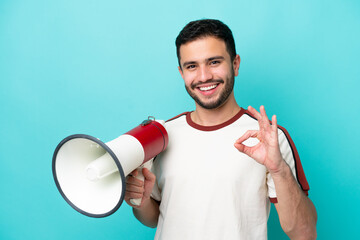 Young Brazilian man isolated on blue background holding a megaphone and showing ok sign with fingers