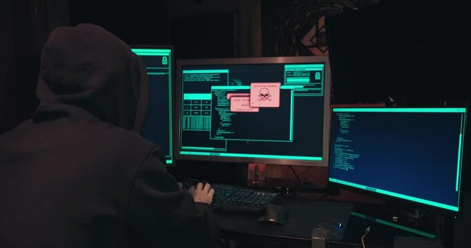 Hacker in a hood sits in front of computer screens and hacks databases. The concept of cyberterrorism, server hacking and cyberattacks. The room is dark, a person is typing code into the command line