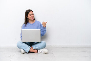 Young caucasian woman with laptop sitting on the floor isolated on white background pointing to the side to present a product