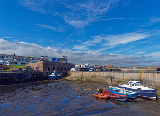 Fototapeta na wymiar North Sunderland harbour at Seahouses at Low Tide, with Boats resting on the silty bottom of the Harbour, overlooked by Houses and Commercial Buildings.