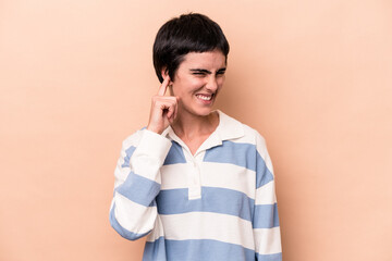 Young caucasian woman isolated on beige background covering ears with fingers, stressed and desperate by a loudly ambient.