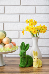 Stand with Easter eggs, rabbit and narcissus flowers on wooden counter near white brick wall