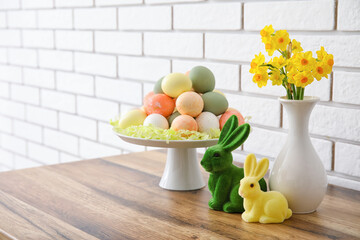 Stand with Easter eggs, rabbit and narcissus flowers on wooden counter near white brick wall