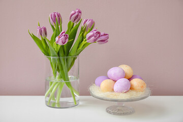 Bouquet of tulips and stand with Easter eggs on table near color wall