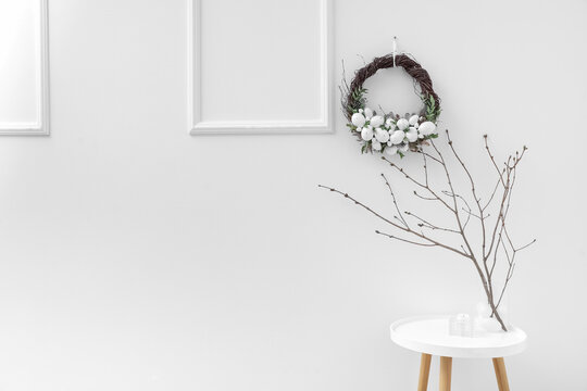 Tree branches in vase on table near white wall with Easter wreath