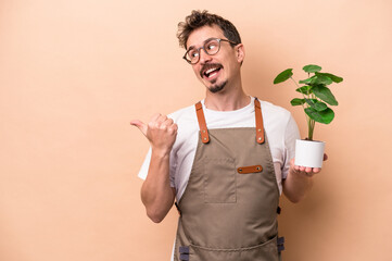 Young caucasian gardener man holding a plant isolated on beige background points with thumb finger...