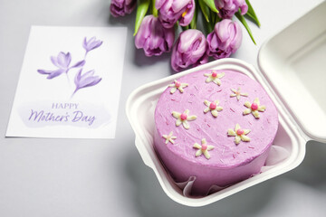 Plastic lunch box with tasty bento cake, greeting card and flowers on grey background, closeup