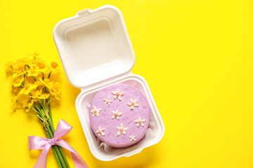 Obraz na płótnie Canvas Plastic lunch box with tasty bento cake and flowers on yellow background
