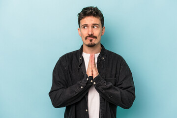 Young caucasian man isolated on blue background praying, showing devotion, religious person looking for divine inspiration.