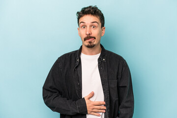 Young caucasian man isolated on blue background touches tummy, smiles gently, eating and satisfaction concept.