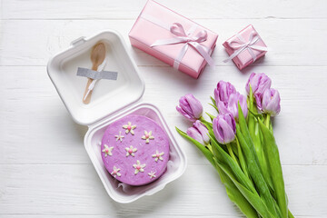 Plastic lunch box with tasty bento cake and flowers on light wooden background