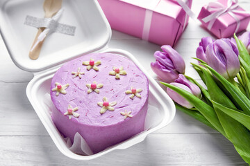 Plastic lunch box with tasty bento cake and flowers on light wooden background, closeup