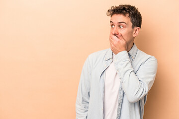 Young caucasian man isolated on beige background thoughtful looking to a copy space covering mouth with hand.