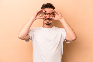 Young caucasian man isolated on beige background showing okay sign over eyes