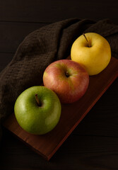 Three multicolored apples on a brown wooden board on a dark brown wooden background.