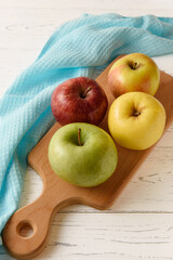A group of four colored apples on a wooden board and a blue napkin on a white wooden background.