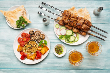 Three servings of appetizing homemade shish kebab, sliced cucumbers and radishes, barbecue vegetables, glasses of wine top view on a blue wooden background.