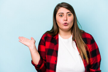 Young caucasian overweight woman isolated on blue background impressed holding copy space on palm.