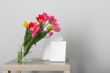 Vase with tulips and blank photo frames on table near light wall