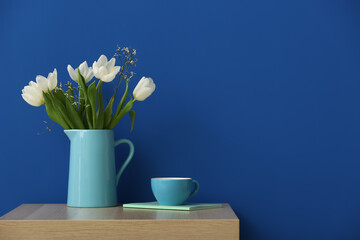 Jug with tulips, cup and notebook on table near color wall