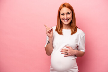 Young caucasian pregnant woman isolated on pink background smiling and pointing aside, showing...