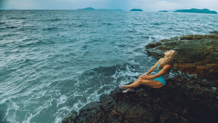Alone attractive woman walk near a rock reef hill in stormy morning rain cloudy sea. Girl in turquoise swimsuit. Concept resort outdoor relax, vacation, loneliness. Dark dramatic view