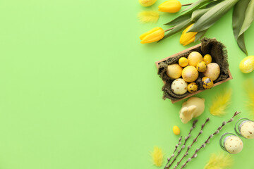 Box with painted Easter eggs, tulip flowers and willow branches on green background