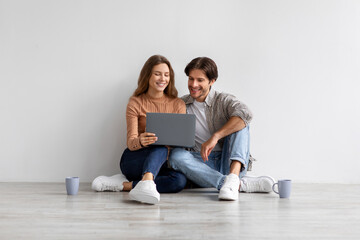 Glad smiling young european man and lady look at laptop planning new interior, sitting on floor in...