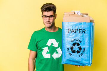 Young caucasian man holding a recycling bag full of paper to recycle isolated on yellow background confused, feels doubtful and unsure.