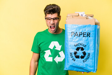 Young caucasian man holding a recycling bag full of paper to recycle isolated on yellow background screaming very angry and aggressive.