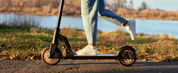 Woman discover city and park at sunset with electric scooter or e-scooter. Female Legs in sports sneakers stand on electric scooter. Girl riding on Ecological and urban transport. © wertinio