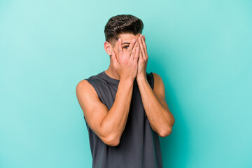 Young caucasian man isolated on blue background blink through fingers frightened and nervous.