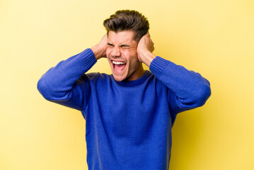 Young caucasian man isolated on yellow background covering ears with hands trying not to hear too loud sound.