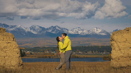 Fototapeta na wymiar Man and woman in yellow green sportswear. Lovely couple of travelers hug and kiss near old stone enjoying highland landscape. Two travelers are walking against the backdrop of snow-capped mountains.