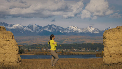 The woman in yellow green sportswear. The traveler near old stone enjoying highland landscape. Tourist are walking against the backdrop of snow-capped mountains. Strong wind.