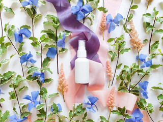 Mockup of unbranded white plastic spray bottle, blue and pink flowers and silk ribbons on white table. Bottle for branding and label. Natural organic spa cosmetics and liquid antimicrobial spray.