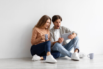 Fototapeta na wymiar Surprised millennial caucasian male and female with open mouths look at tablet sit on floor in empty room