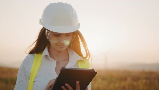 Caucasian woman smiling and looking at camera with digital tablet in hands. Female person wearing formal clothes, helmet and glasses on windmill farm. Concept of people, technology and green energy