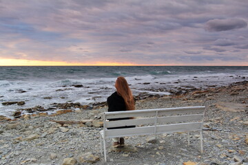 a lone figure of a woman from behind on a bench on a rocky seashore during sunset