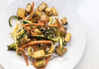Tofu recipe with vegetables, carrot, zucchini, red and green pepper with asian noodles on a white plate