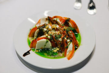 A portion of caprese on a white plate on a white table. A traditional Italian starter of mozzarella cheese, with fresh tomatoes, pesto sauce and balsamic sauce.
