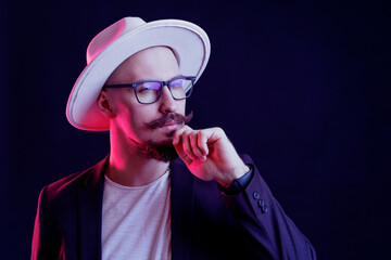 Sceptic bearded man in white wide-brimmed hat in blazer in glasses holds fist on chin. Color filters
