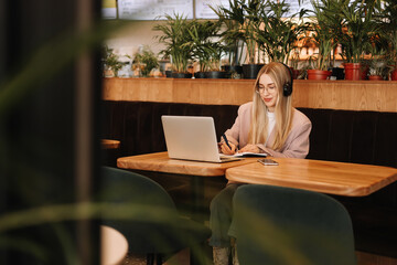 A young millennial freelancer woman with headphones works and studies online using a mobile phone and wireless technologies while sitting in the workplace. A business woman takes notes in a planner