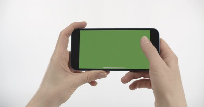 Close-up of Smartphone with Green Screen in Woman's Hands. Scrolling the Phone Screen Up. Smartphone with Green Screen in Horizontal Position on White Background