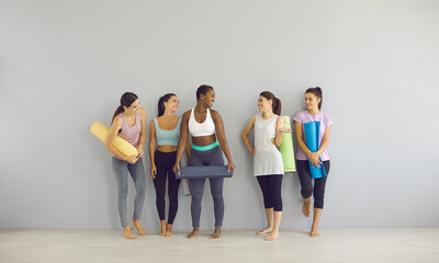 Group of smiling girls in modern sports outfits and with workout mats standing in fitness studio....