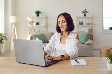 Beautiful smiling business lady working remotely in her home office using laptop. Middle-aged woman sitting at table in front of laptop and notebook and looking for useful information on Internet.