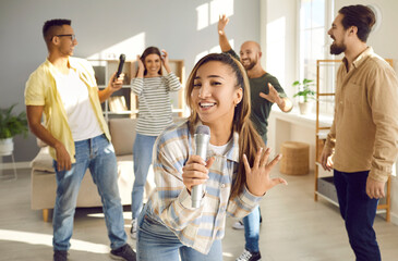 Positive young woman holding microphone and singing karaoke during home party with friends. Woman singing song while her cheerful friends are dancing and having fun in background. Fun concept.